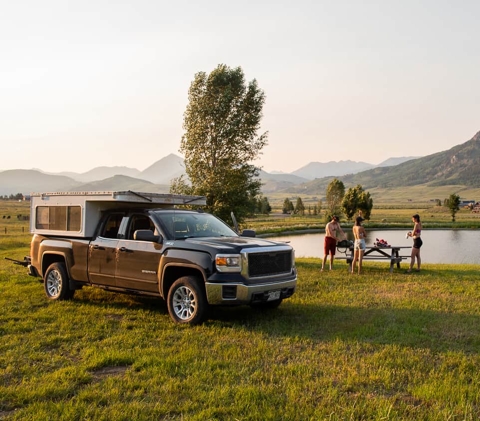 Pickup truck camper parked on a lakeside meadow with mountains behind