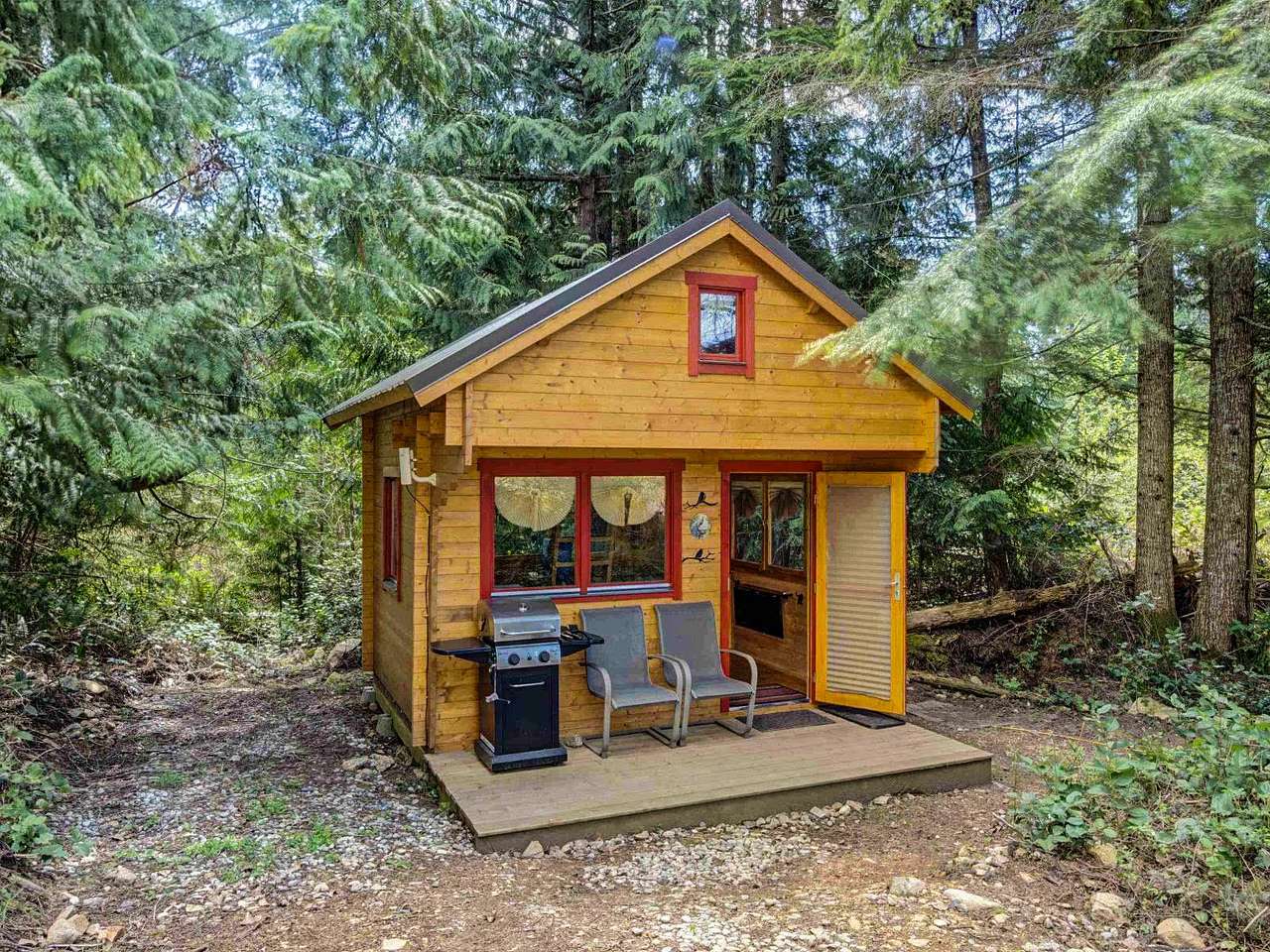 The Peacock cabin is nestled in the woods. The front deck features a barbecue and two comfy chairs. There is a separate area on the same site with an outdoor dining table.